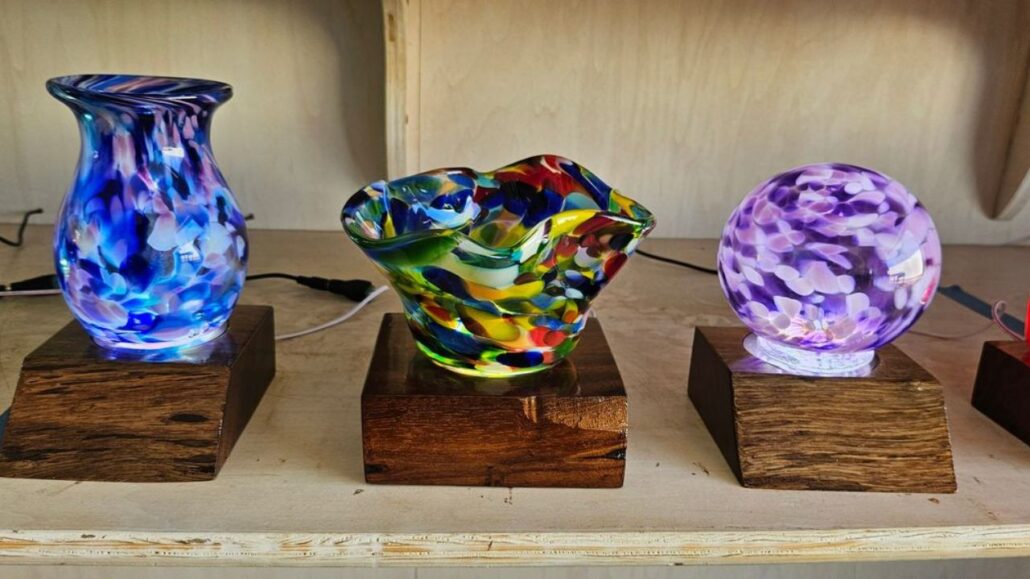 Colorful hand-blown glass ornaments displayed on wooden lighted bases, including a blue vase, a multicolor bowl, and a purple orb with soft glowing illumination.