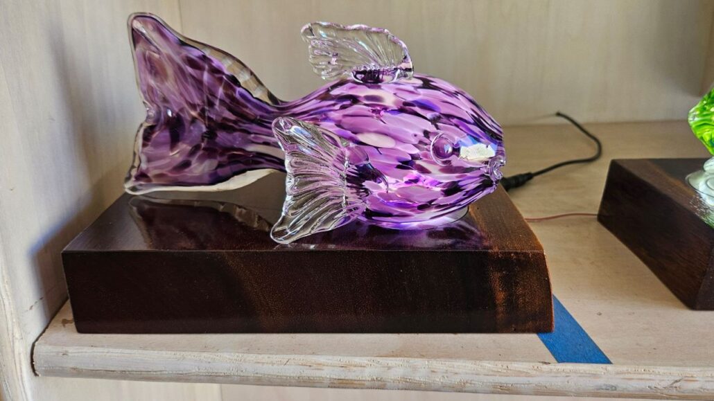 Purple glass fish sculpture displayed on a wooden lighted base at Moana Glass.
