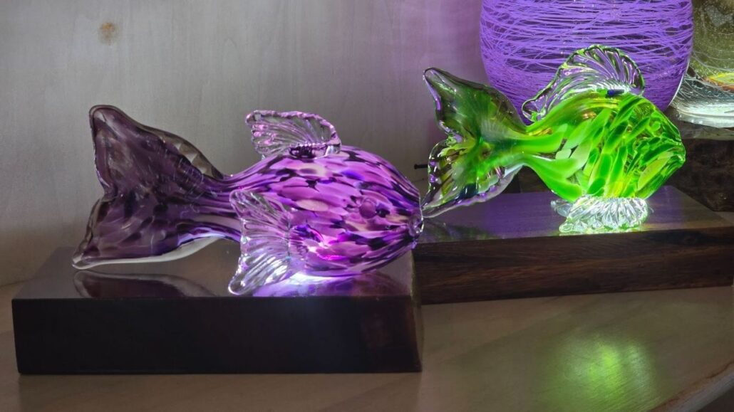 Handmade glass art pieces on wooden bases, including colorful sculpted glass fish displayed on a studio shelf.