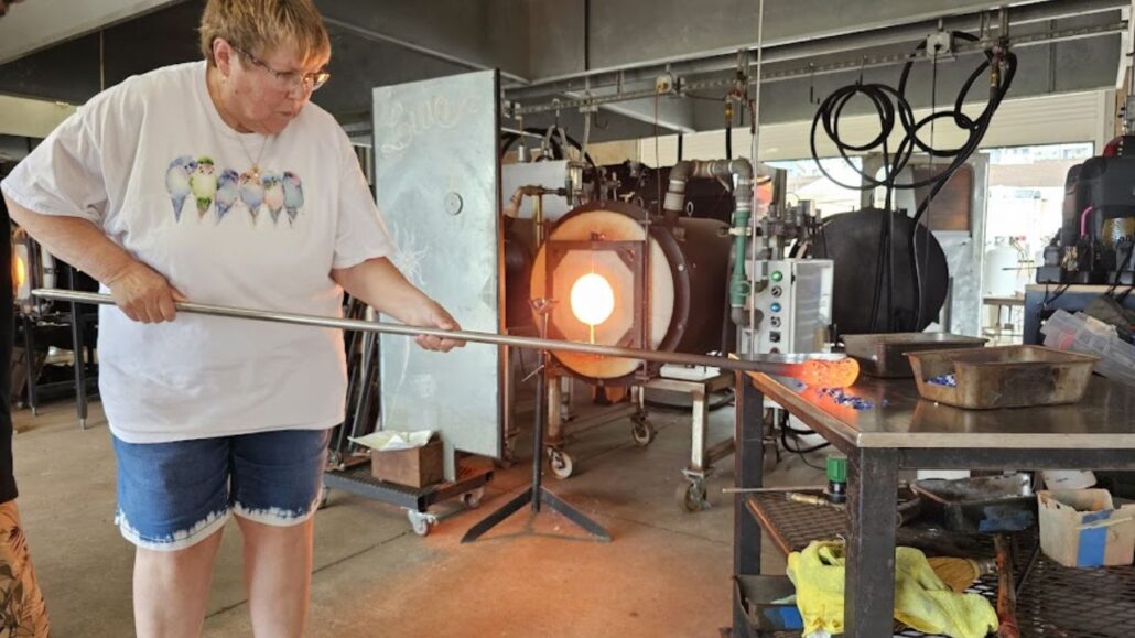 Participant gathers molten glass from a glowing furnace using a blowpipe in a glassblowing studio.